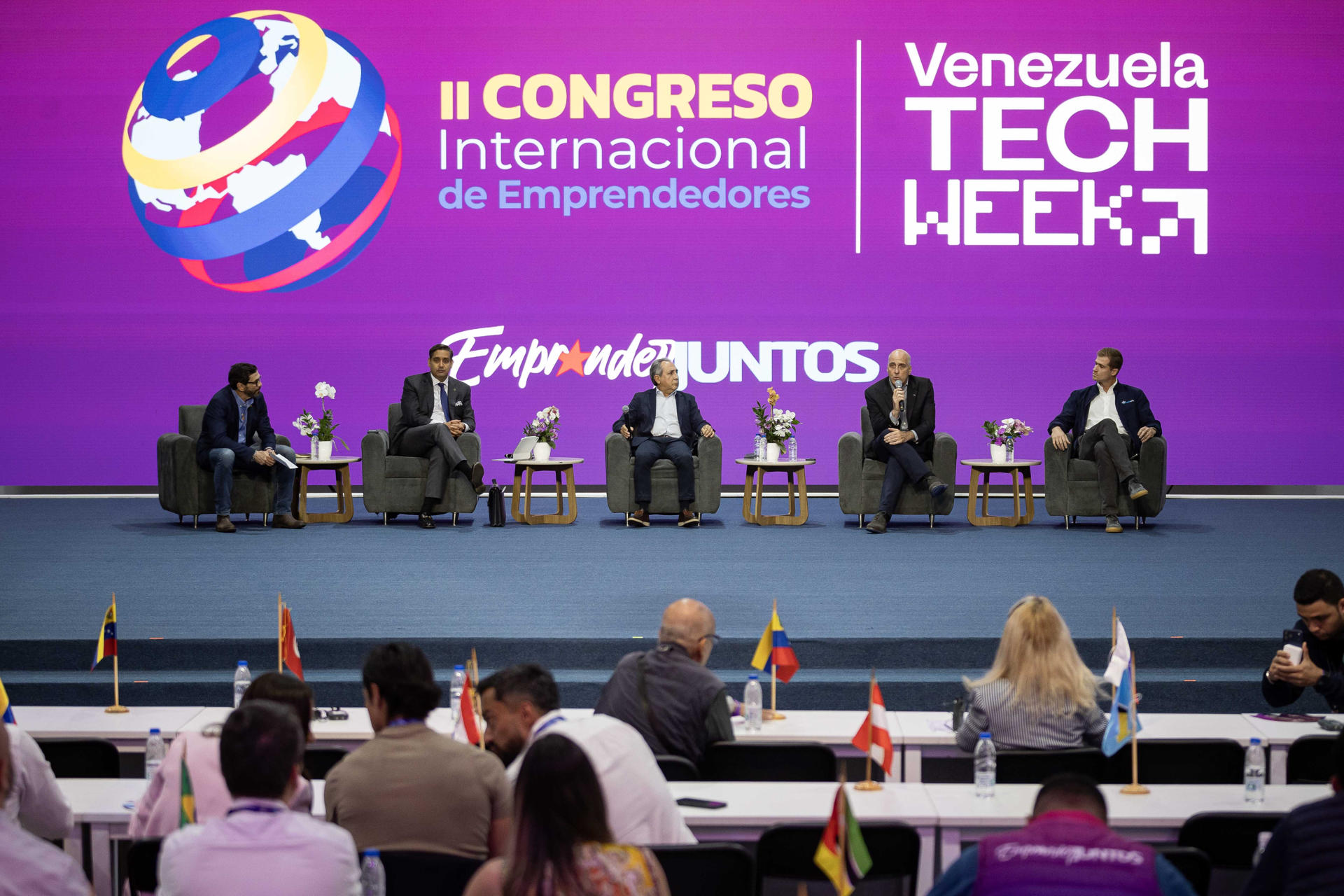 Un grupo de empresarios venezolanos conversando en un evento de tecnología, con logos de empresas visibles y un ambiente de negocios.