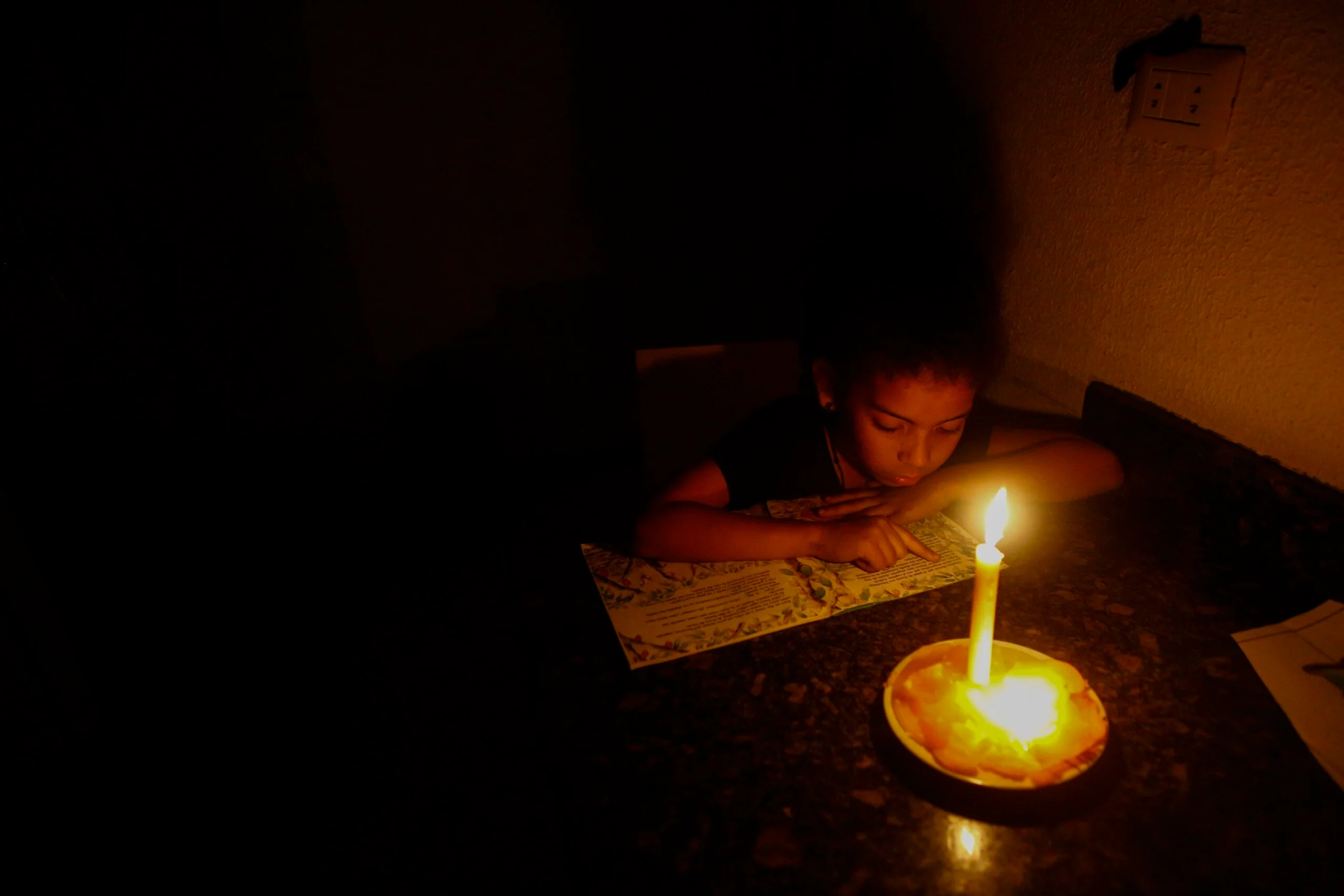 Niños venezolanos en un salón de clases con poca luz, sentados en pupitres, mientras uno intenta escribir.