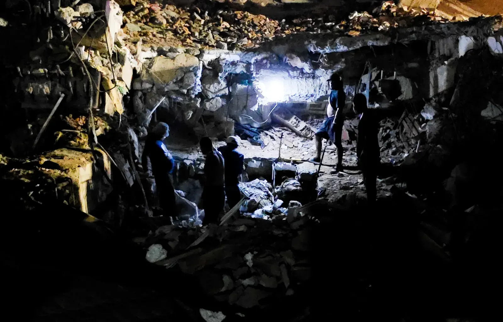 damnificados en La Guaira reunidos en una acera iluminados por lámparas recargables, con escombros y edificios dañados al fondo.